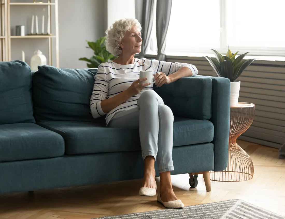 A smiling senior woman holding a cup of tea beside a sunlit window